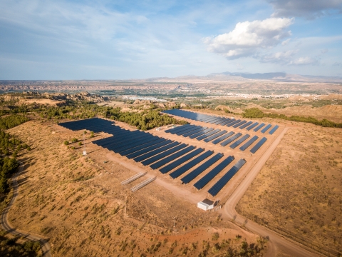 This aerial photograph captures rows of dark blue solar panels that stretch across a sun-baked desert landscape under a cloudy sky. The setting includes a dusty desert terrain and rolling hills in the background. The photo highlights sustainable power and environmental awareness. This scenic photo is suitable for projects focusing on renewable energy, conservation, and modern technology. The image evokes a sense of calm, showcasing the harmony between technology and the natural world.