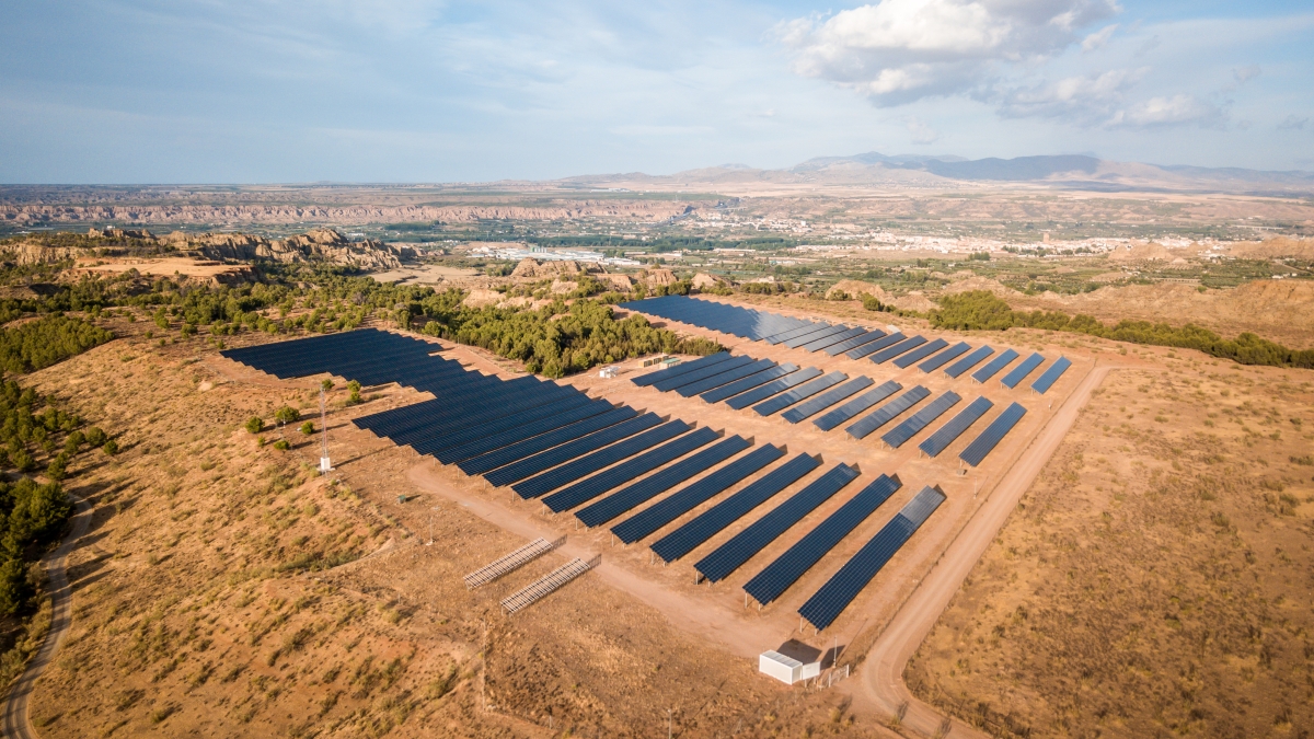 This aerial photograph captures rows of dark blue solar panels that stretch across a sun-baked desert landscape under a cloudy sky. The setting includes a dusty desert terrain and rolling hills in the background. The photo highlights sustainable power and environmental awareness. This scenic photo is suitable for projects focusing on renewable energy, conservation, and modern technology. The image evokes a sense of calm, showcasing the harmony between technology and the natural world.